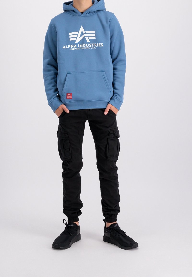 Felpa con cappuccio blu con logo bianco "Alpha Industries", abbinata a pantaloni cargo neri con più tasche e sneakers nere.