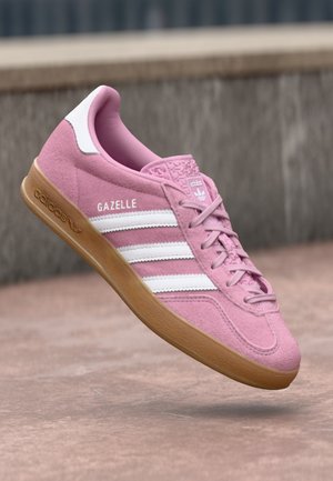 Baskets Adidas Gazelle roses avec des rayures blanches et une semelle gomme, vues de côté contre un fond flou de béton en plein air.
