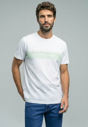 Hombre con cabello rizado y barba, vestido con camiseta blanca estampada y vaqueros azules, de pie frente a un fondo gris.