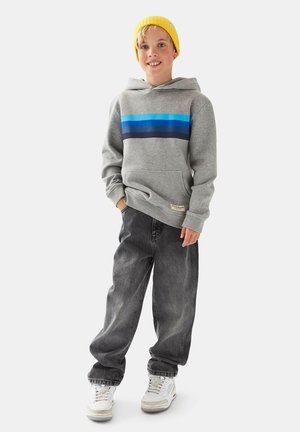 Smilende barn iført gul hue, grå hættetrøje med blå striber, løse sorte jeans og hvide sneakers, stående foran en ensfarvet baggrund.