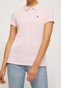 Žena v krátkém rukávu světle růžového polo trička s malou tmavě modrou golfer logem a světle modrými džíny, na jednobarevném pozadí.