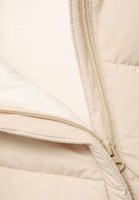 Bežová prešívaná bunda s hladkým povrchom, mäkkou fleece podšívkou a svetlým detailom zipsu, ktorý čiastočne odhaľuje vnútornú textúru.