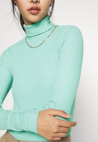 Turtleneck-Oberteil in Mintgrün, aus geripptem Stoff gefertigt, mit langen Ärmeln und slim fit. Inklusive einer dünnen goldenen Kettenhalskette als Akzent.