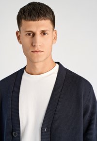 Granatowy prążkowany kardigan z głębokim dekoltem w kształcie litery V i zapięciem na guziki, noszony na białej koszulce. Gładka faktura, nowoczesny design, dopasowana sylwetka.