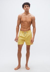 Νεαρός άνδρας με σγουρά μαλλιά στέκεται ξυπόλυτος φορώντας κίτρινα μαγιό σε απλό λευκό φόντο.