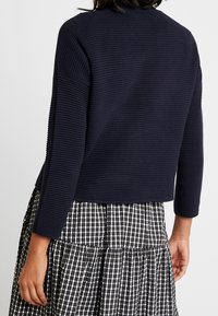 Granatowy sweter z prążkowanej dzianiny z rękawami o długości trzech czwartych; w połączeniu z czarno-białą spódnicą w kratę z tierowanymi falbanami.