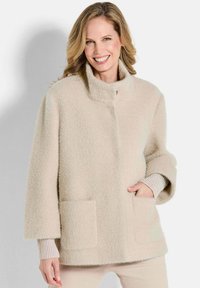 Beige gek textureerde jas met een staande kraag, twee voorkruiken en brede mouwen, gelaagd over een geribbelde lichte trui.