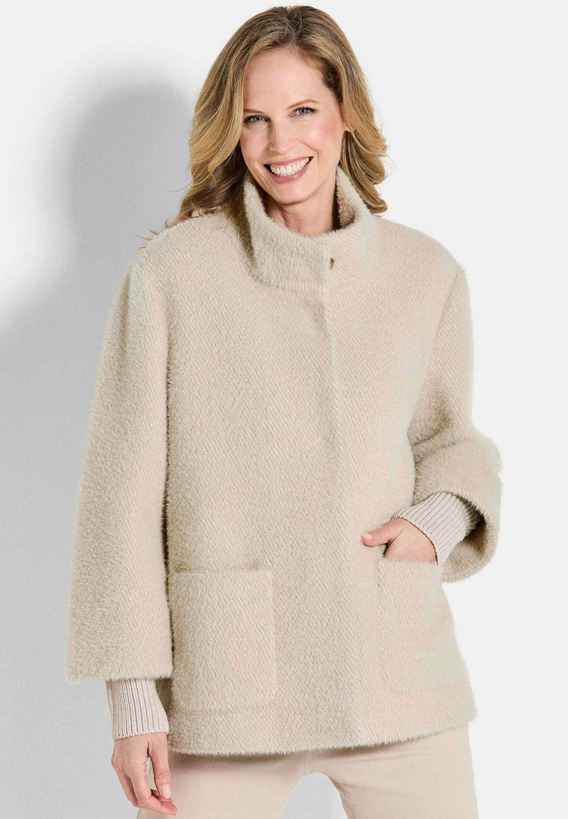 Beige gek textureerde jas met een staande kraag, twee voorkruiken en brede mouwen, gelaagd over een geribbelde lichte trui.