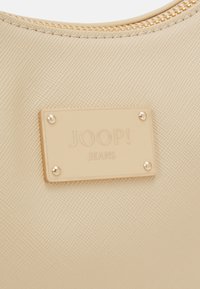 Beżowy materiał teksturowany z złotym zamkiem. Złota tabliczka z napisem "JOOP! JEANS" ozdobiona czterema dekoracyjnymi nitami.