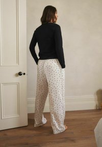 Woman standing barefoot on wooden floor, wearing black long sleeve top and white wide-leg pants with black polka dots, facing away indoors.