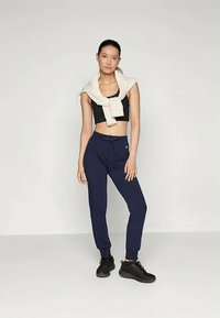 Sieviete stāv ar melnu īso krekla augšu, tumši ziliem sporta biksēm, melnajām kedām un balto džemperi pāri pleciem pret vienkāršu fona.