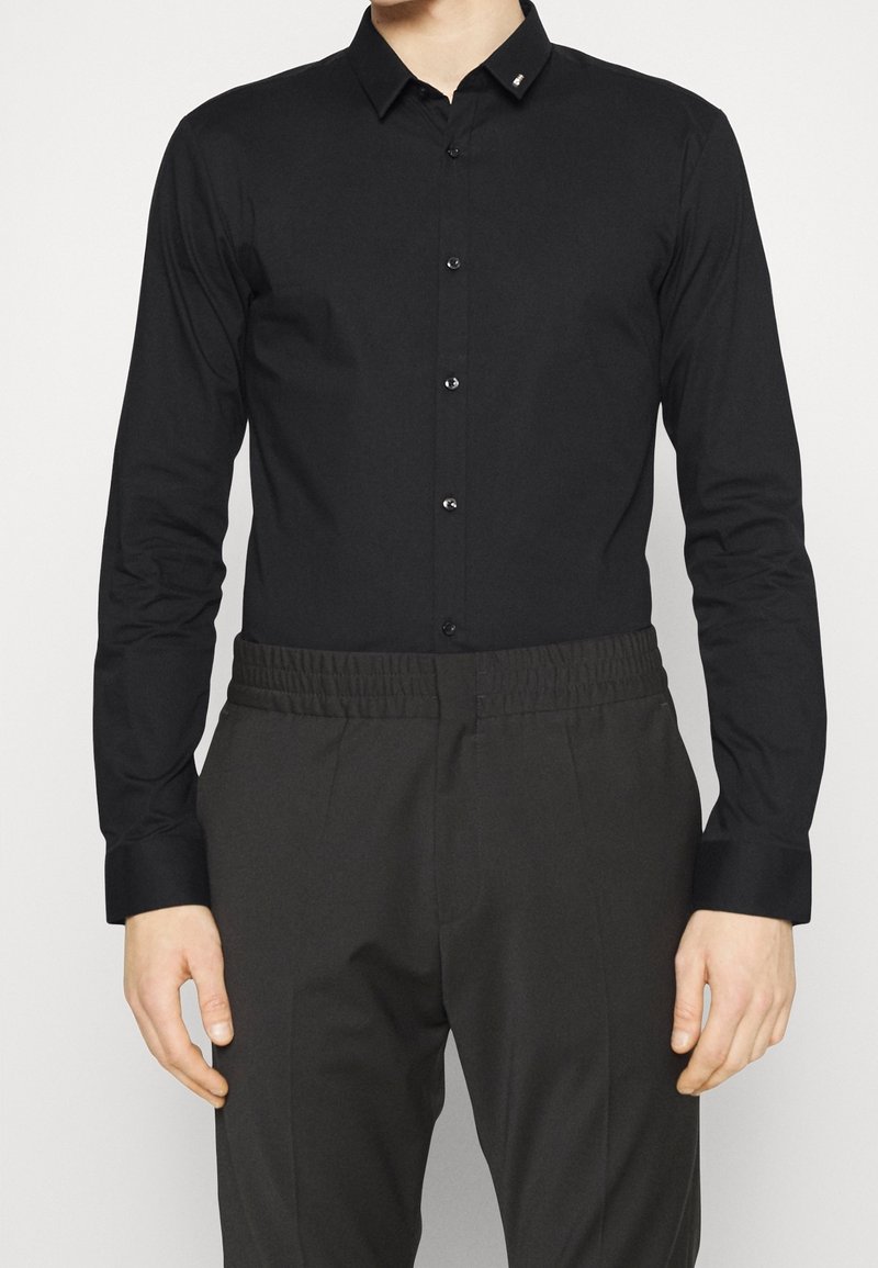 Homme portant une chemise noire cintrée à manches longues boutonnée, rentrée dans un pantalon noir à taille élastique, debout devant un fond uni.