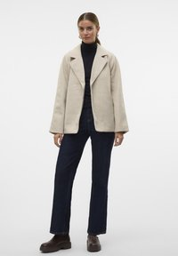 Giacca beige in misto lana con colletto a scollo, apertura frontale e maniche ampie, abbinata a jeans in denim scuro e stivali alla caviglia marroni.