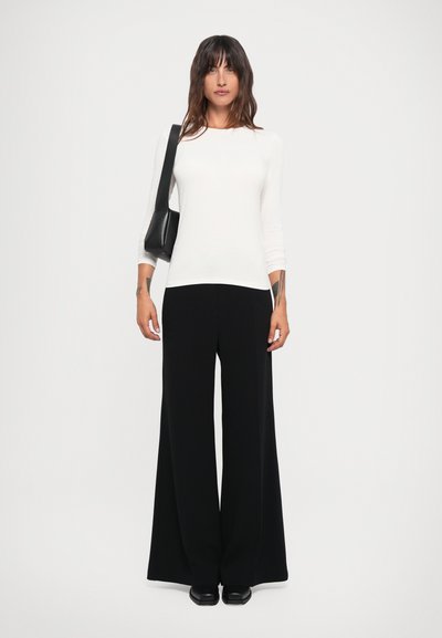 Haut à manches longues blanc avec un col rond, associé à un pantalon large noir. Sac à épaule noir supplémentaire et chaussures noires épaisses visibles.