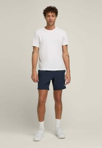 Camiseta blanca de cuello redondo combinada con pantalones cortos azul marino. El modelo lleva deportivas blancas y calcetines. Diseño sencillo y casual con un ajuste relajado.