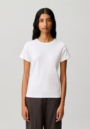 Donna con i capelli lunghi e neri indossa una semplice t-shirt bianca a maniche corte e pantaloni scuri a righe sottili, in piedi su uno sfondo grigio chiaro.