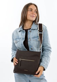 Vrouw met licht denimjack en spijkerbroek, die een donkerbruine crossbodytas van getextureerd leer met een voorvak met rits vasthoudt.