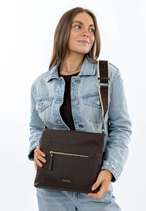 Vrouw met licht denimjack en spijkerbroek, die een donkerbruine crossbodytas van getextureerd leer met een voorvak met rits vasthoudt.