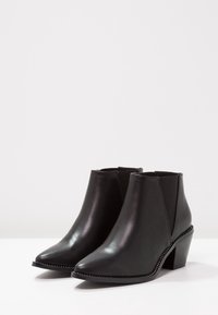 Černé kožené kotníkové boty s špičatou špičkou, elastickými bočními panely a patou se stacked heels. Obsahují dekorativní perlový detail podél podrážky.