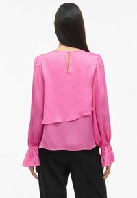 Rosa bluse med lange, rynkede ermer, lagdelt bakdesign og en nøkkelhullåpning ved halsen. Laget av glatt, flytende stoff.