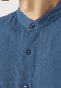 Primer plano de una camisa de lino azul con cuello mao y tres botones negros, mostrando el área del cuello y la parte superior del pecho.