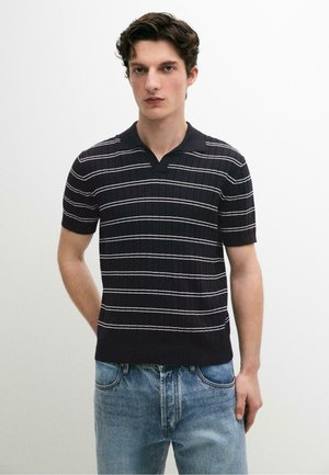 Giovane uomo con capelli corti e scuri che indossa una polo nera a maniche corte con strisce orizzontali bianche e jeans chiari, in piedi su sfondo bianco.