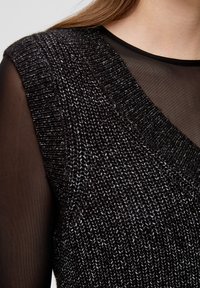 Černý a stříbrný pletený bezrukávový svetr nošený přes průsvitný černý dlouhý rukáv, odhalující klíční kost a oblast ramen.