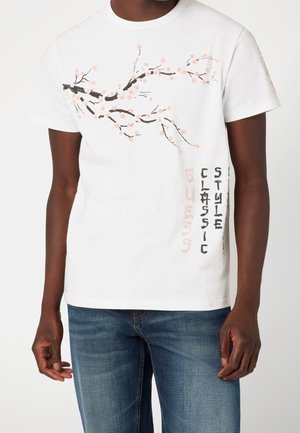 Man wears white short-sleeve T-shirt with black and pink cherry blossom branch and vertical "classic style" text, paired with blue jeans.