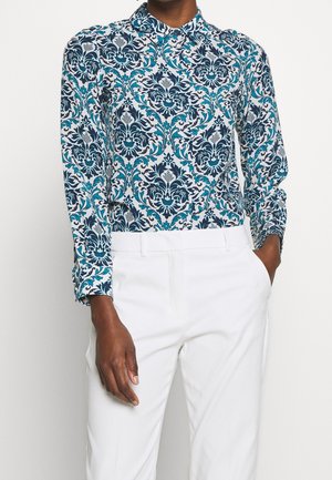 Kobieta ubrana w koszulę z długim rękawem i kołnierzykiem w niebiesko-turkusowy wzór kwiatowy, włożoną do białych spodni z wysokim stanem.