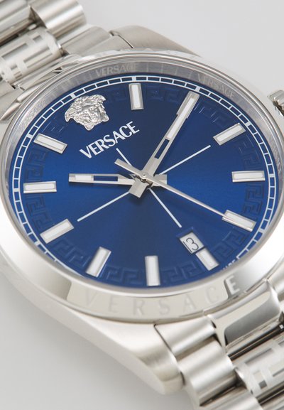 Montre en acier inoxydable argenté avec un cadran bleu, présentant un logo de tête de lion, des index horaires géométriques et un affichage de la date à trois heures.