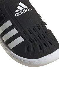 Svart treningssko med teksturert overflate, hvitt tre-stripes design og perforeringer for ventilasjon. Har en hvit gummisåle.