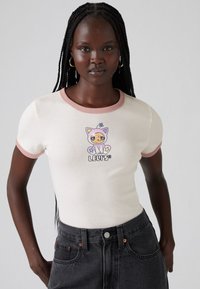 Kvinne med lange fletter har på seg en hvit ringer-t-skjorte med rosa kant og en fargerik katt-grafikk, kombinert med høytidte svarte jeans.