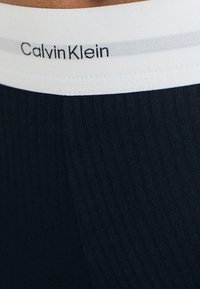 Crni elastični pojas s natpisom "Calvin Klein" u bijelom tekstu i svijetlosivom prugom. Tkanina ima rebrastu teksturu.