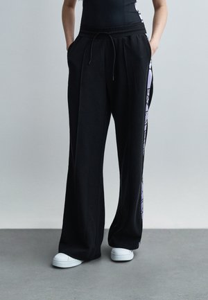 Osoba oblečená v čiernych širokých teplákoch s bočnými pruhmi a bielych teniskách, stojaca s rukami v vreckách na sivom pozadí.