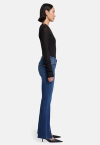 Czarna bluzka z długim rękawem, niebieskie dżinsy z szerokimi nogawkami i postrzępionym dołem oraz czarne szpilki z ostrym noskiem. Widok z boku ukazuje dopasowaną sylwetkę i tekstury.