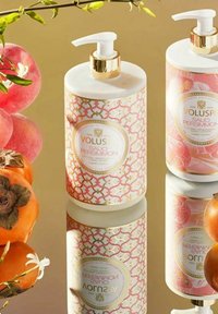 Voluspa håndlotion og skrub i hvide cylindriske flasker med pink geometriske mønstre og guld accenter, omgivet af persimmoner og æbler.