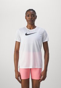 Balts Nike sporta krekls ar melnu svītru, kuram ir rozā gradiente apakšmala. Apvienots ar rozā šortiem, nodrošinot pieguļošu izskatu un mīkstu tekstūru.
