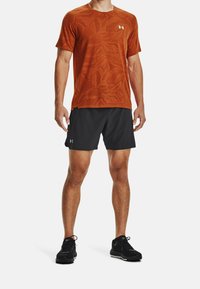 Oranžové sportovní tričko s krátkým rukávem a listovým vzorem, doplněné černými kraťasy. Černé běžecké boty se šněrovadly dokončují outfit.