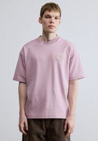 Svetloružové bavlnené tričko s krátkymi rukávmi a okrúhlym výstrihom. Má dekoratívnu nášivku na ľavej strane hrudníka. Hladká textúra.