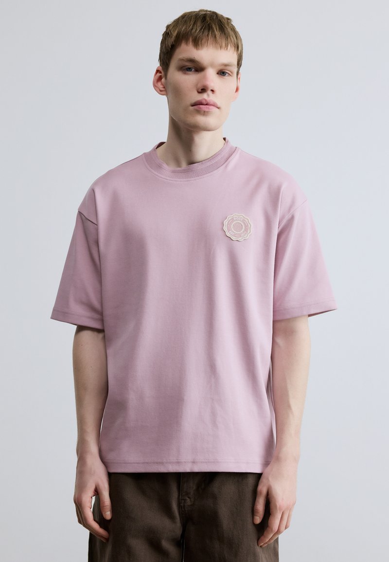 Svetloružové bavlnené tričko s krátkymi rukávmi a okrúhlym výstrihom. Má dekoratívnu nášivku na ľavej strane hrudníka. Hladká textúra.