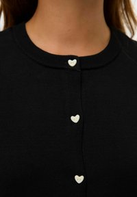 Gros plan sur un cardigan noir avec de petits boutons en forme de cœur blancs le long du devant, près de l'encolure.