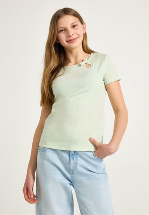 Ung kvinde med langt hår iført en lysegrøn, ribstrikket kortærmet top med udskæringsdetaljer og lyseblå denimjeans, poserer med hånden i lommen.