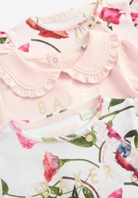 Tres camisetas de tres capas para niños en rosa y blanco, con estampados florales, cuellos rizados y detalles del logo dorados. Textura de tela suave.
