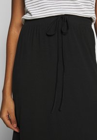 Jupe noire à cordon de serrage avec une texture douce et fluide. La ceinture présente un design froncé avec un lien ajustable. La main du mannequin montre l'ajustement.