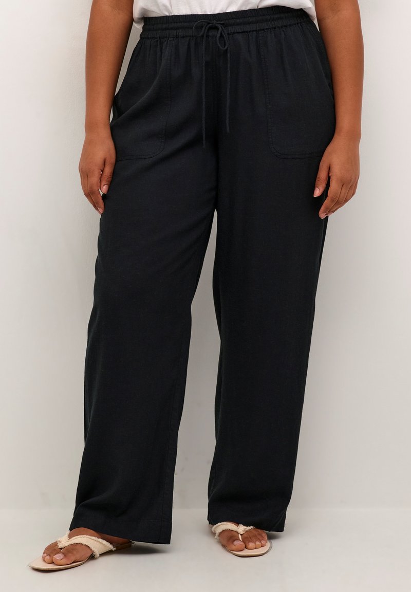 Pantalones anchos negros, confeccionados en una tela suave con una cinturilla elástica, que cuentan con bolsillos laterales y un ajuste relajado. Llevados con sandalias ligeras.