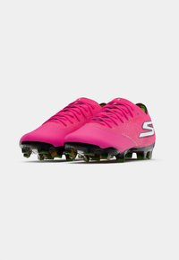 Botas de fútbol de color rosa brillante con suelas y tacos negros, con un logo blanco de "S" en el lateral, colocadas una al lado de la otra sobre un fondo liso.