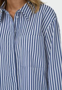 Blau-weiße vertikal gestreifte Button-Up-Bluse mit Kragen und einer aufgesetzten Tasche auf der linken Seite, im entspannten Schnitt und mit weichem Material.