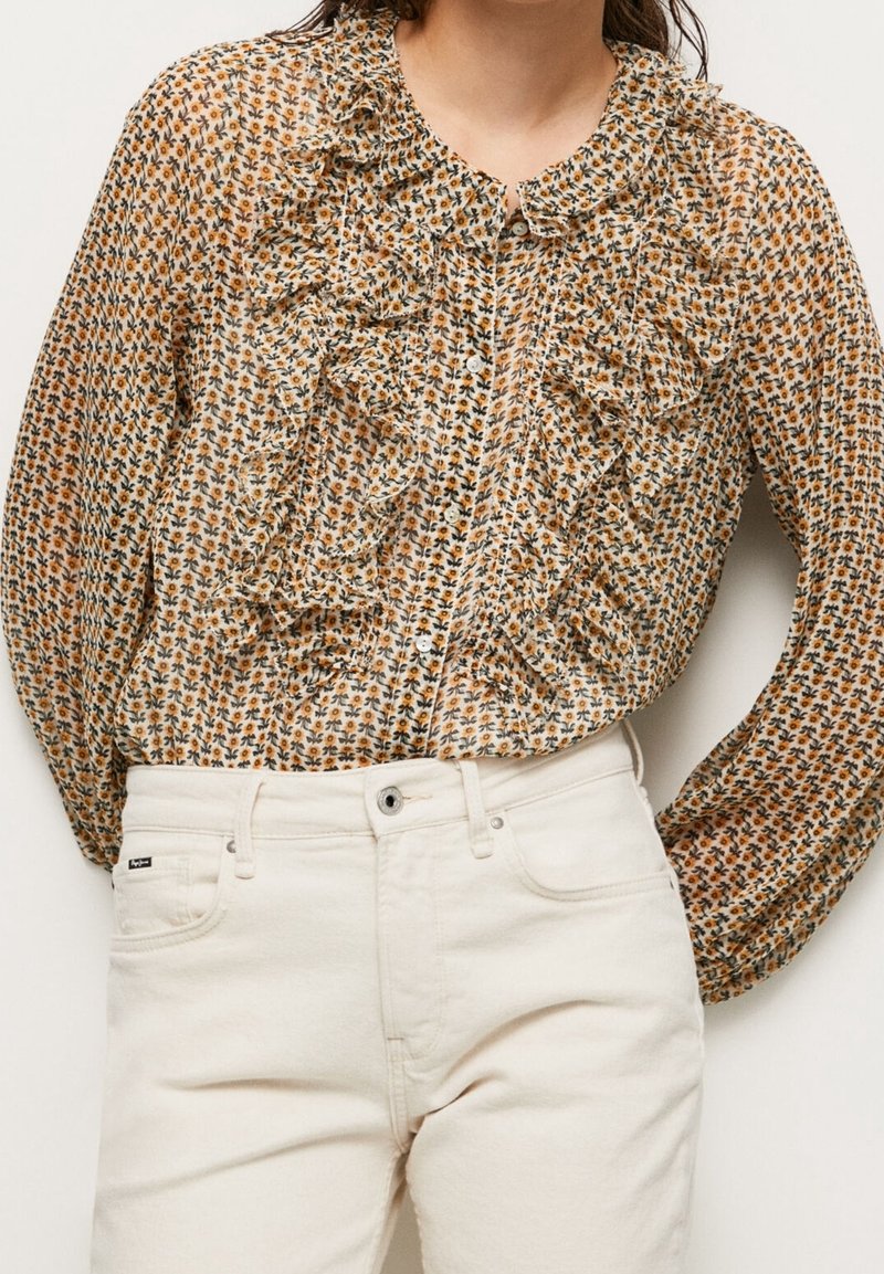 Femme portant un chemisier fleuri à manches longues avec volants, assorti à un pantalon blanc taille haute sur fond uni.
