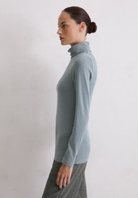 Frau mit braunen Haaren in einem Dutt, die einen hellblauen Rollkragenpullover und graue Hosen trägt, seitlich vor einer schlichten weißen Wand stehend.