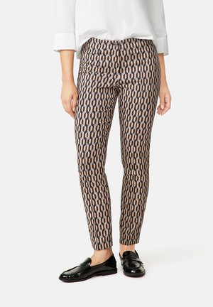 Femme portant un pantalon slim à motifs noir et beige avec un chemisier blanc et des mocassins noirs brillants, debout devant un fond uni.